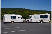 Two white shuttle vans parked facing each other in an empty parking lot with tree-covered hills and a clear blue sky behind them.