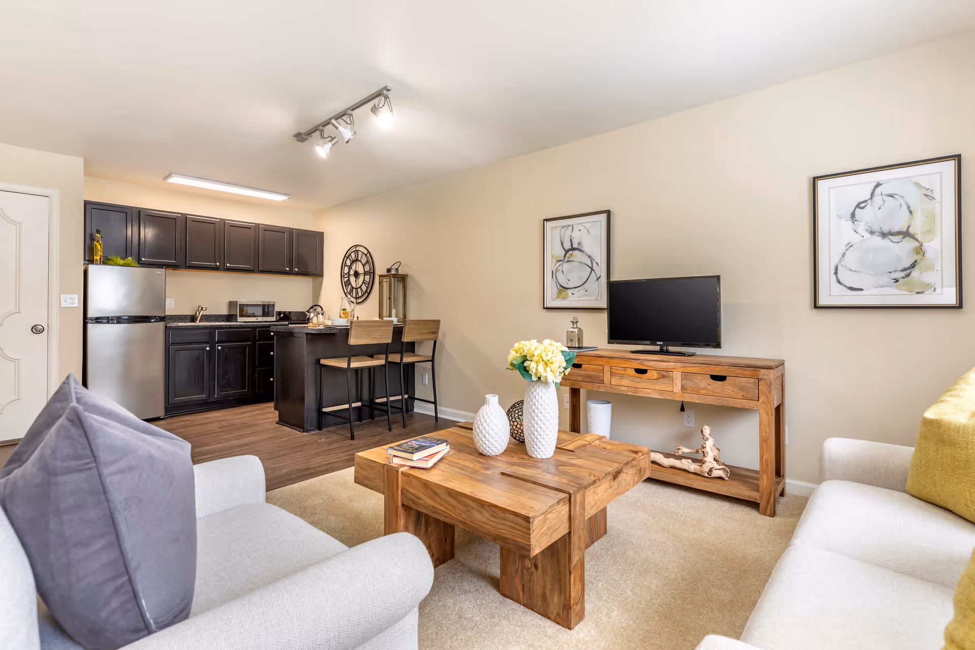 A cozy living room and kitchen area with beige walls and carpet. The living room has two light-colored sofas with gray and yellow pillows, a wooden coffee table with decorative vases and books, and a wooden TV stand with a flat-screen TV and two framed abstract paintings on the wall. The kitchen features dark cabinets, a stainless steel refrigerator, a microwave, a sink, and a small breakfast bar with two wooden stools. The room is well-lit with ceiling lights.