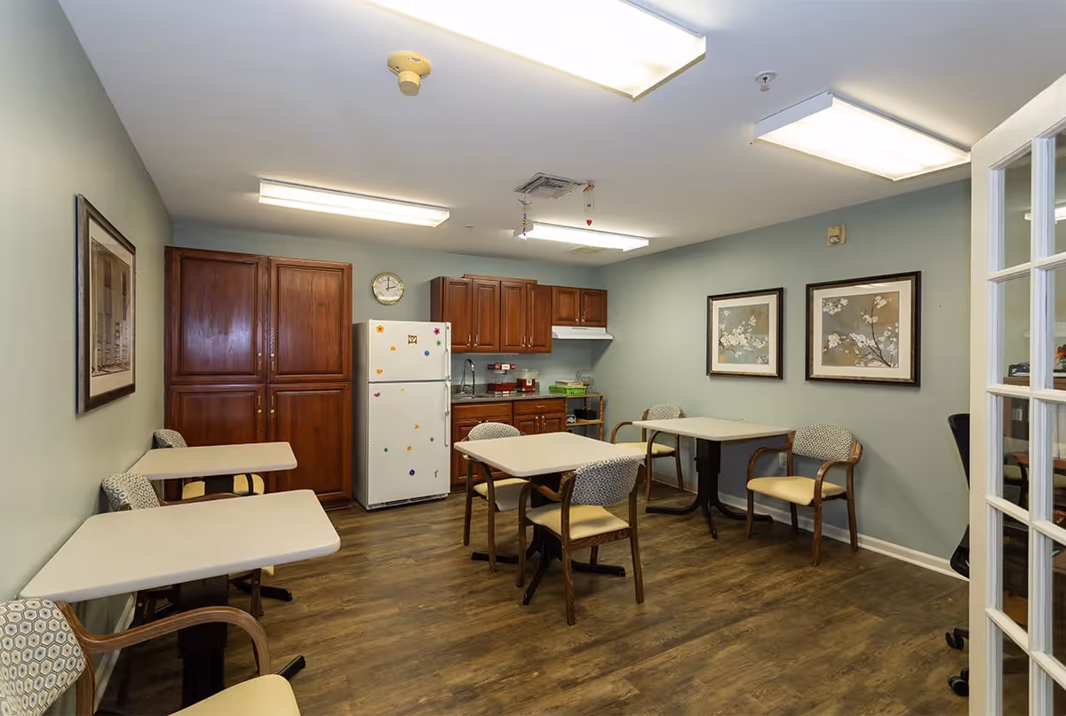 A small dining area with four tables and several chairs, a white refrigerator decorated with colorful magnets, wooden cabinets, a sink, and two framed floral pictures on the wall. The room has wood flooring and fluorescent ceiling lights.