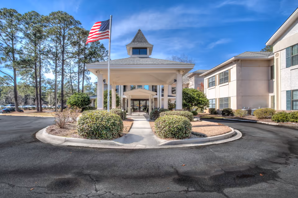 Front entrance of a multi-story senior living building with a covered porte-cochère, American flag, driveway, and landscaped shrubs.