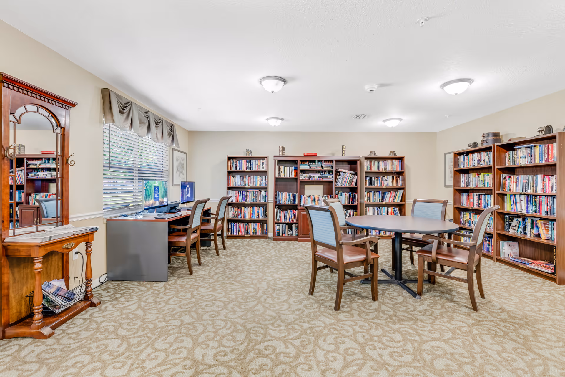 A well-lit room with beige walls and patterned carpet featuring several wooden bookshelves filled with books. There is a round table with four chairs in the center of the room, and a desk with two computers and chairs along one wall under a window with blinds and a valance. A wooden hall tree with a mirror and a small shelf is positioned near the desk.