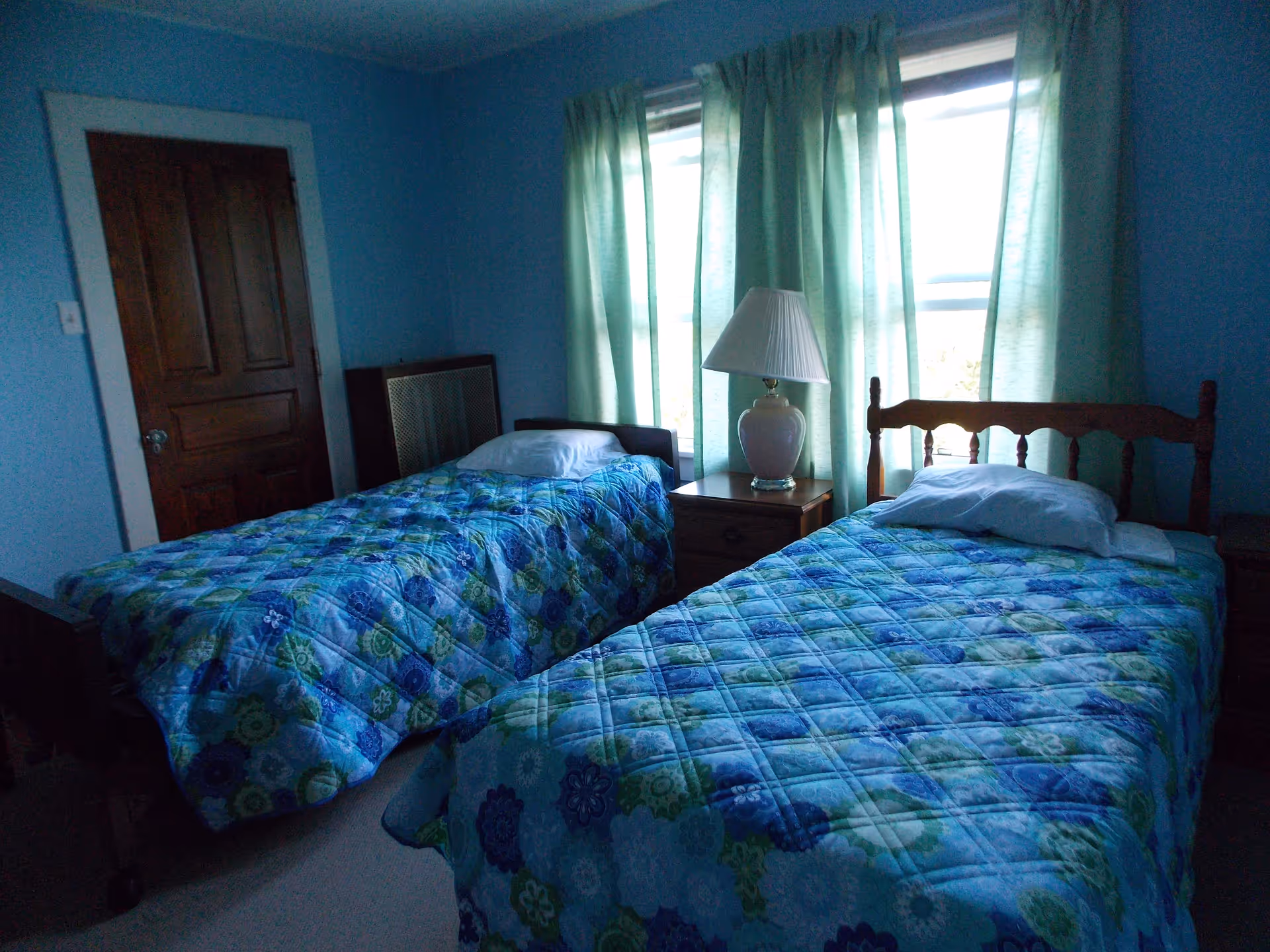 A bedroom with two single beds covered with blue and green floral patterned quilts. Each bed has a white pillow. Between the beds is a wooden nightstand with a white lamp. The room has light blue walls, a wooden door, and two windows with light green curtains.