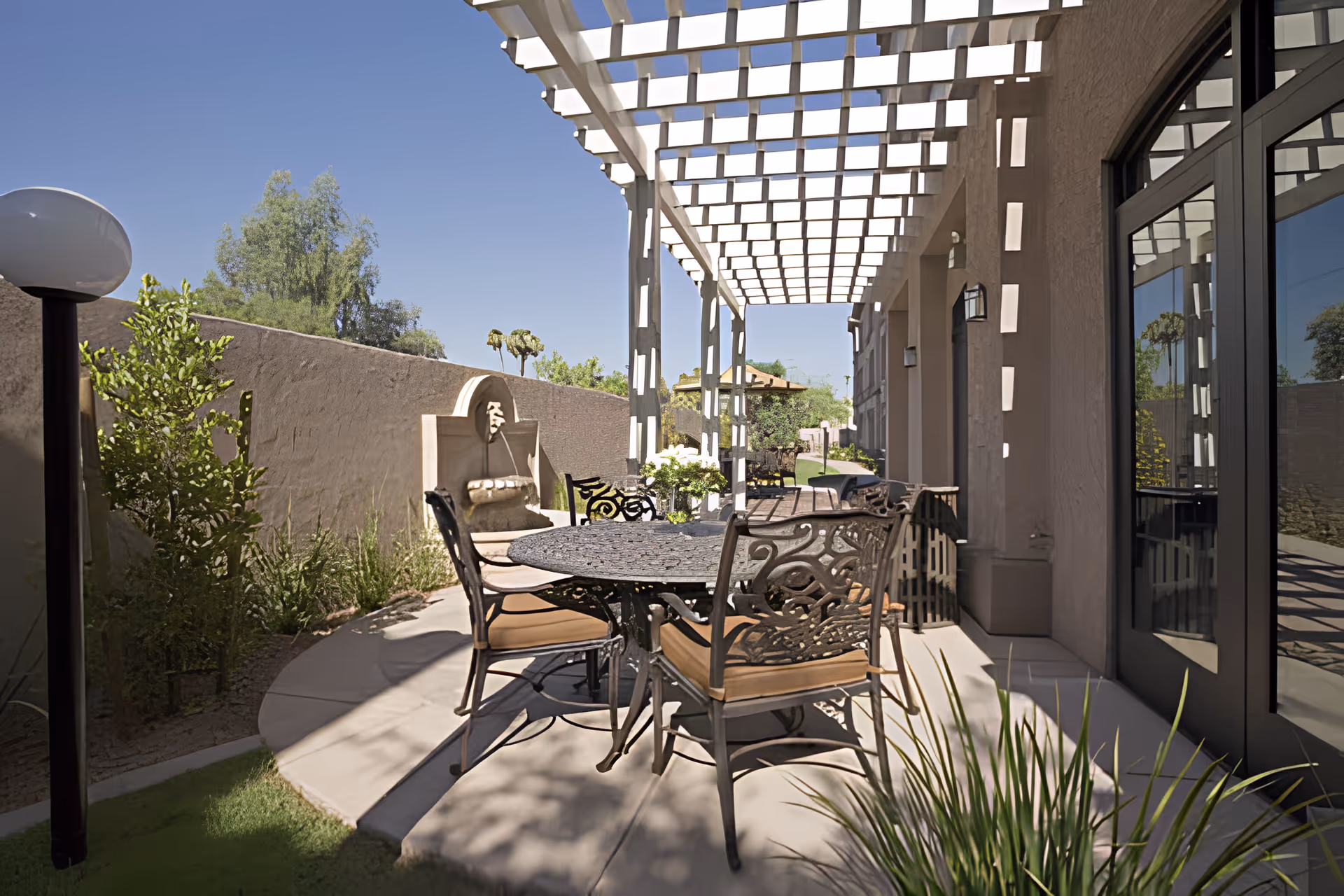 Outdoor patio seating area with a round metal table and chairs under a white pergola beside a beige building and a small fountain.
