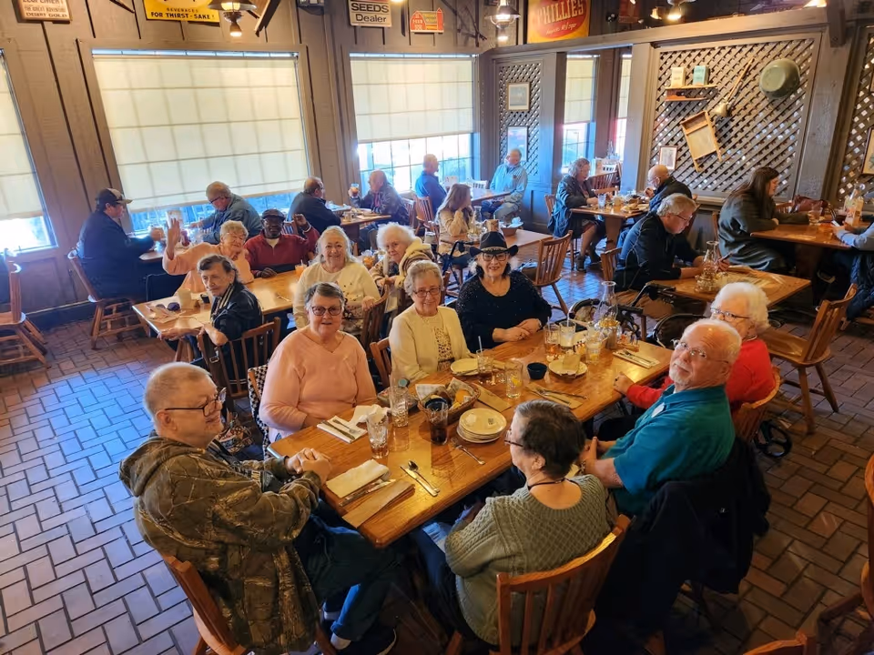 A group of elderly people sitting around wooden tables in a cozy dining room with brick flooring and large windows covered by blinds. The room has rustic decor with vintage signs and wooden lattice panels on the walls. The people appear to be enjoying a meal together, with plates, glasses, and utensils on the tables.