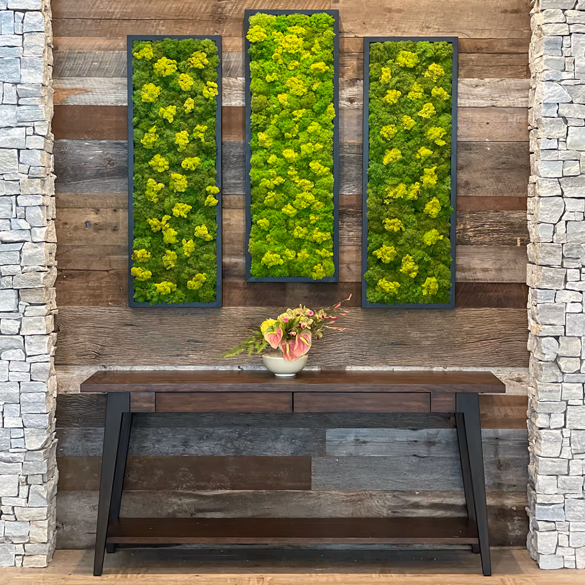 Wood-paneled interior wall with three vertical green moss art panels above a wooden console table holding a small floral arrangement, flanked by stone columns.