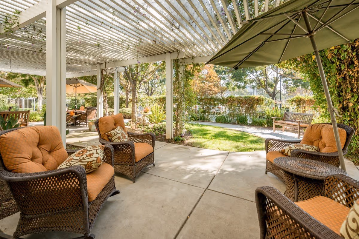Outdoor patio area with cushioned wicker chairs and a table under a pergola, surrounded by greenery and trees with a lawn and garden in the background.