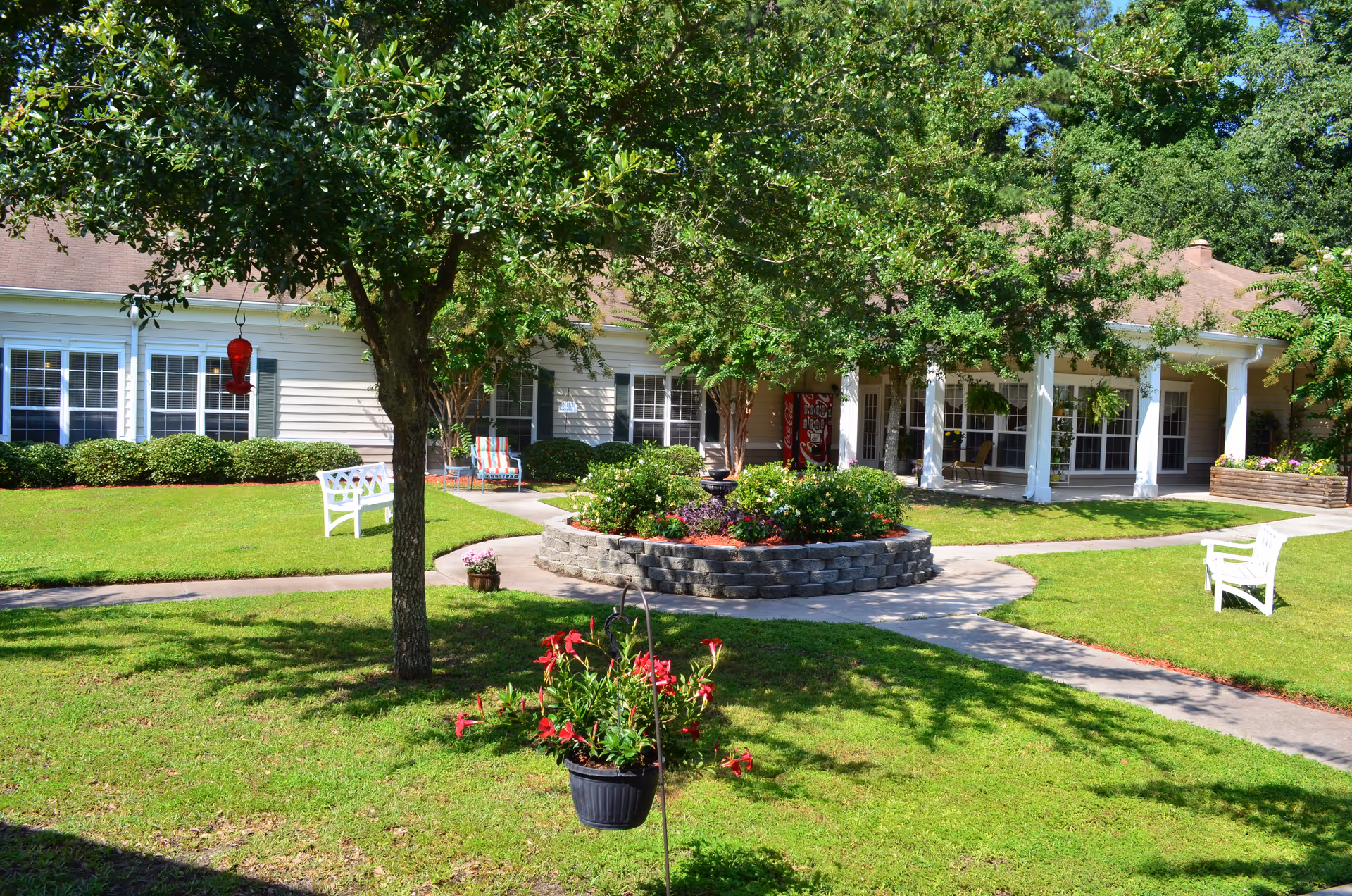 A sunny outdoor garden area at Oaks At Salem Road featuring a large tree, green grass, flower beds, white benches, and a building with large windows and a covered porch in the background.