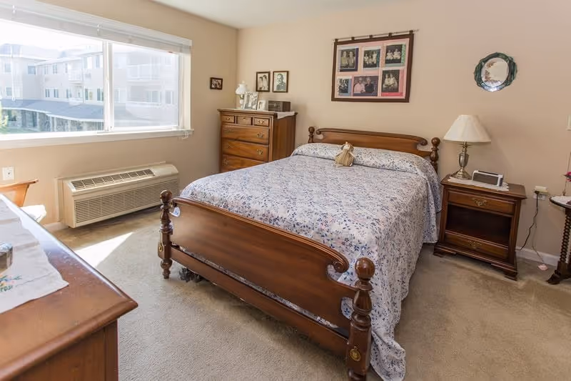 A cozy bedroom in Creekside Oaks Retirement Community featuring a wooden bed with a floral bedspread, a wooden dresser and chest of drawers, a nightstand with a lamp and clock, framed photos on the walls, and a large window letting in natural light.