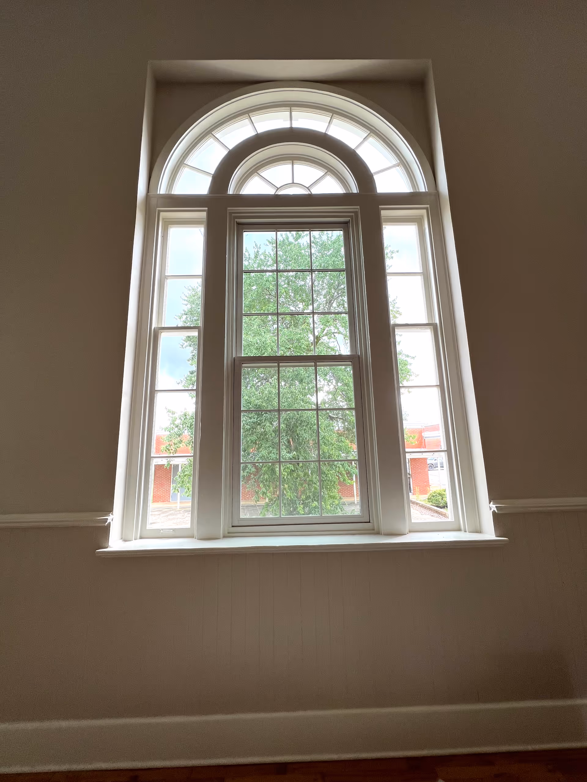 Tall interior wall featuring a large arched multi-pane window overlooking a tree and a neighboring brick building.