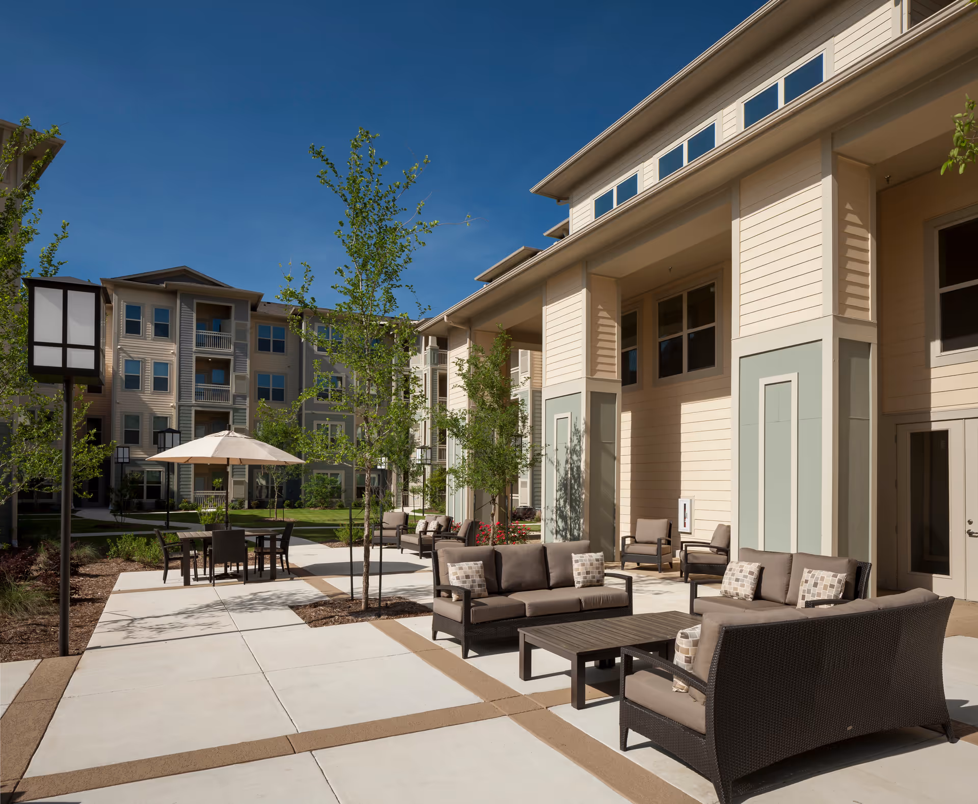 Outdoor patio area at Discovery Village Stone Oak with modern wicker seating including sofas and chairs with cushions, a table with an umbrella, young trees, and a multi-story residential building in the background under a clear blue sky.