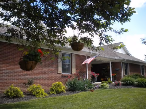 Exterior view of a single-story brick building with a porch, an American flag hanging near the entrance, hanging flower baskets, and well-maintained landscaping with green shrubs and a lawn.