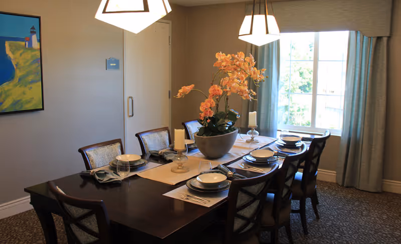 A formal dining room with a dark wood table set for eight, a central floral arrangement, pendant lights, and a window with curtains.