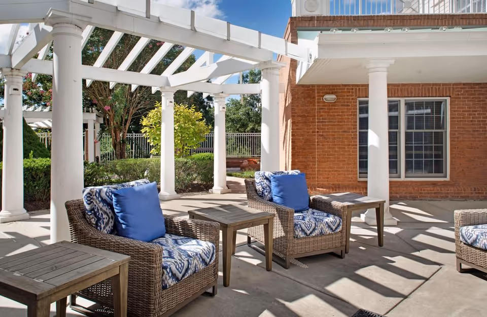 Outdoor patio area with wicker chairs featuring blue and patterned cushions, wooden side tables, white columns, and a pergola structure. The background includes green bushes, trees, and a brick building with windows.