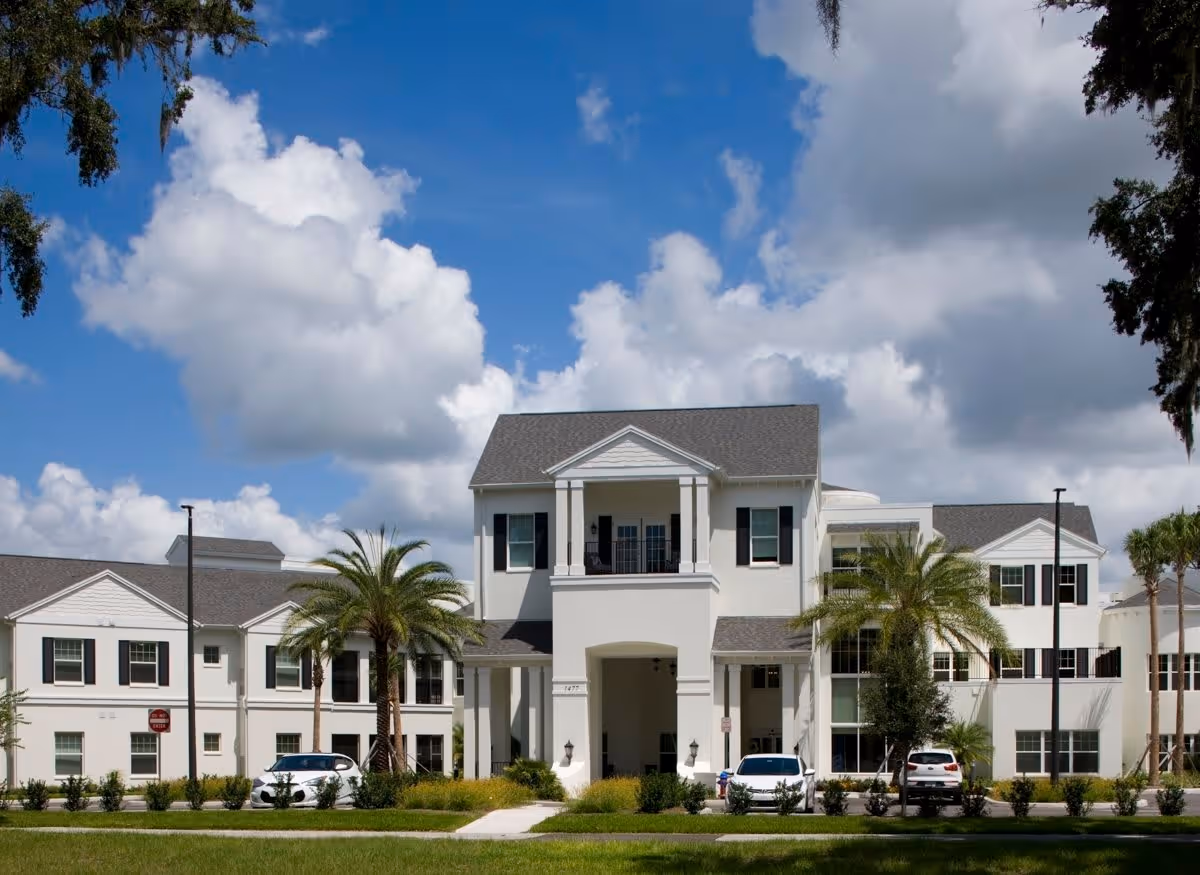 Front exterior view of a large, white senior living facility building with multiple windows, a covered entrance with columns, palm trees, and parked cars under a partly cloudy blue sky.