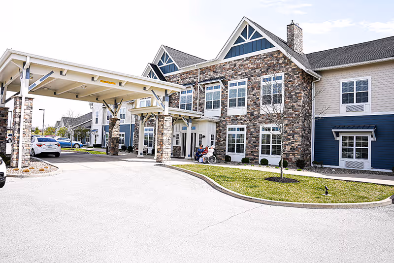 Exterior view of Traditions at Hunter Station, showing a large building with stone and blue siding, multiple windows, and a covered entrance. Two people are sitting near the entrance, one in a wheelchair. Several cars are parked nearby under a clear sky.