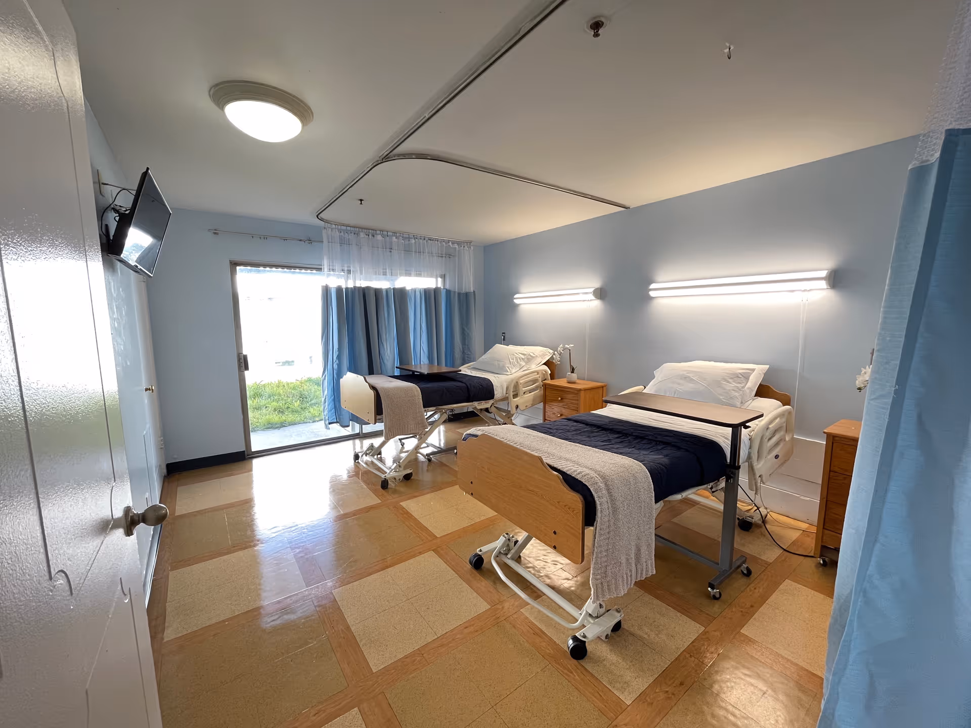 A hospital-style room with two adjustable beds, each with a navy blue blanket and a beige throw. Each bed has a wooden bedside table and an overbed table. The room has light blue walls, a tiled floor with a checkered pattern, and a large window with blue curtains letting in natural light. A wall-mounted TV is visible on the left side.