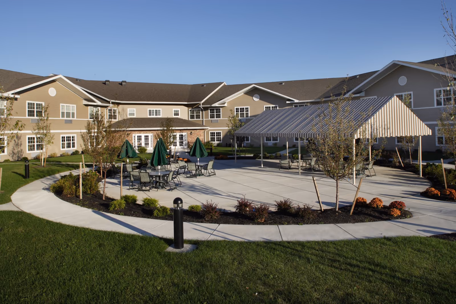 Outdoor patio area at Elderwood Residences at Wheatfield with tables, chairs, and green umbrellas, surrounded by a circular walkway, small trees, and landscaped plants, with a large beige building in the background under a clear blue sky.
