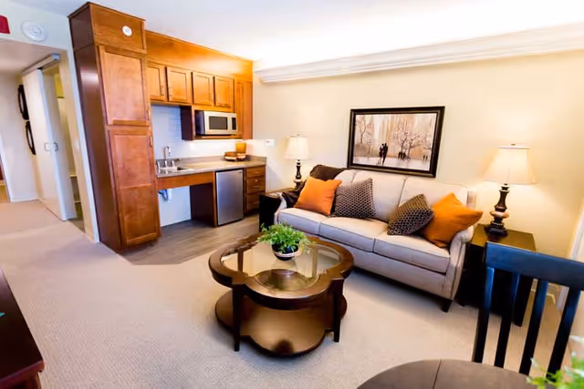 A cozy living room area in a senior living facility featuring a beige sofa with orange and patterned throw pillows, two side tables with lamps, a framed artwork on the wall, a glass-top coffee table with a small plant, and a kitchenette with wooden cabinets, a microwave, a small sink, and a mini refrigerator.