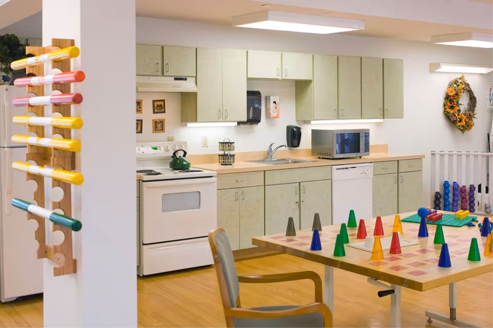A well-lit kitchen area with light green cabinets, a white stove, dishwasher, microwave, and sink. A wooden table with colorful game pieces is in the foreground, along with a chair. A rack holding colorful cylindrical objects is mounted on a white pillar to the left. A decorative wreath hangs on the wall to the right.