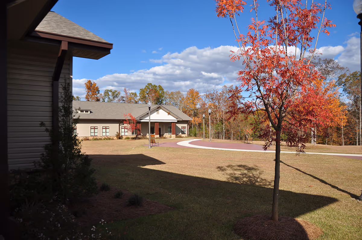 Outdoor view of a senior living facility with a paved walkway, a small tree with red leaves, and a building with a covered entrance. The sky is partly cloudy and there are trees with autumn foliage in the background.