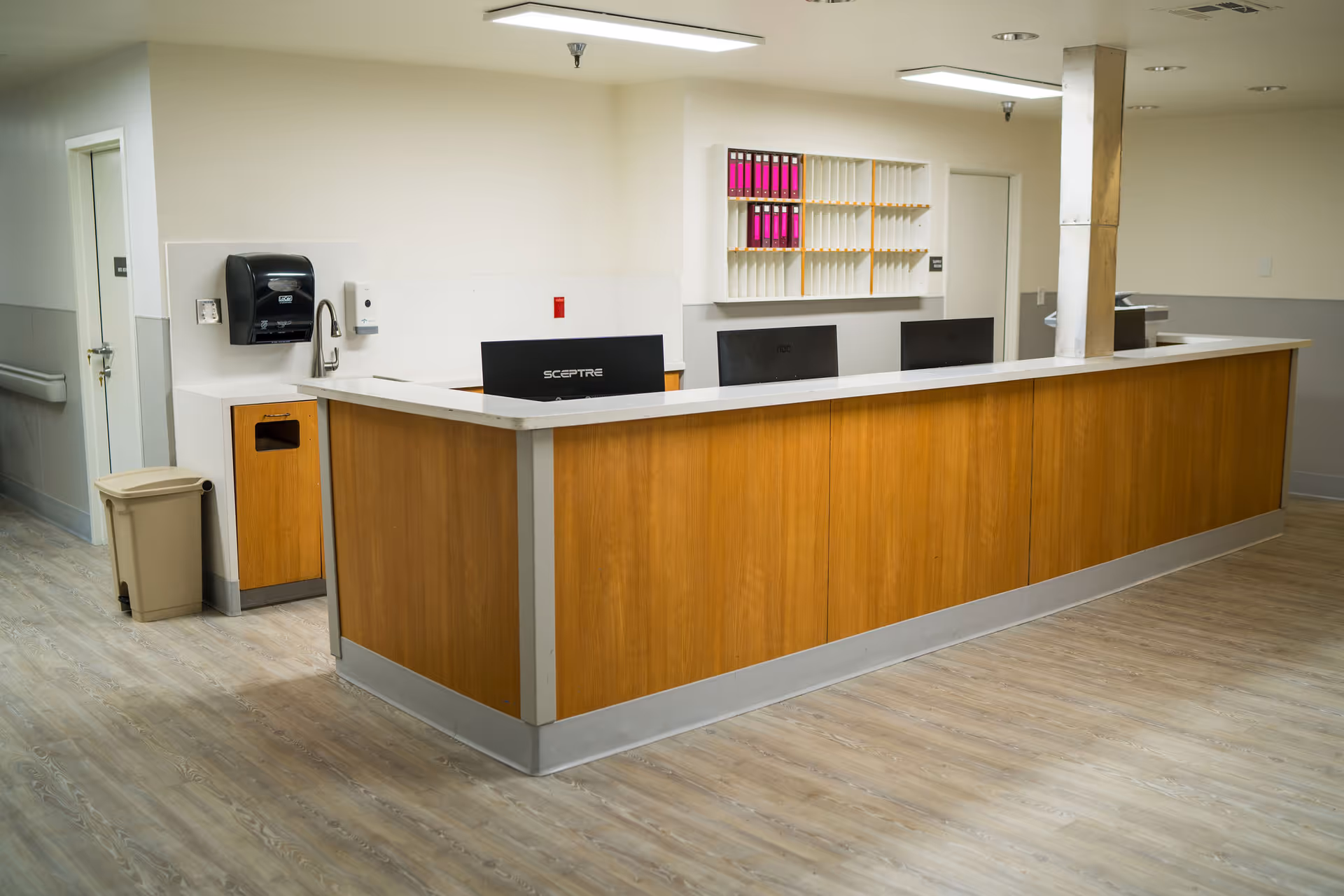 A clean and modern reception desk area in a healthcare facility with wooden paneling and a white countertop. Behind the desk are computer monitors, a wall-mounted organizer with pink folders, and two doors. The floor has light wood-style flooring, and there is a trash bin and a wall-mounted paper towel dispenser on the left side.