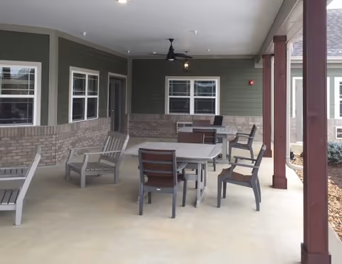 Covered outdoor patio area with several gray tables and chairs arranged for seating. The patio has a concrete floor, green siding with brick accents on the walls, and windows. There are red wooden support beams and a ceiling fan overhead.