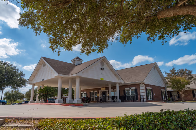 Front entrance of a single-story brick nursing facility with a covered porte-cochere, white columns, and landscaping under a blue sky.