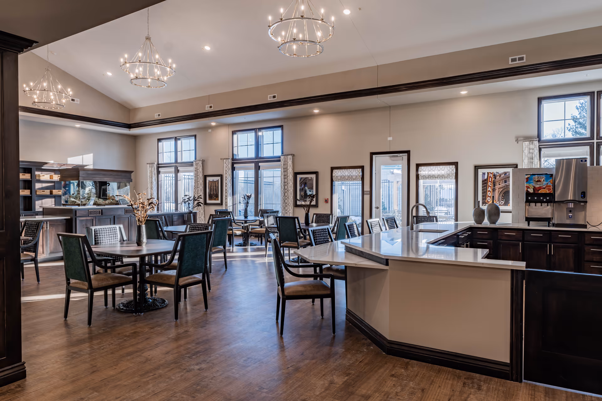 A spacious and well-lit dining area in a senior living facility featuring multiple tables and chairs arranged neatly on a wooden floor. The room has large windows with curtains allowing natural light to enter, modern chandeliers hanging from a high ceiling, and a counter area with a beverage dispenser and decorative vases.