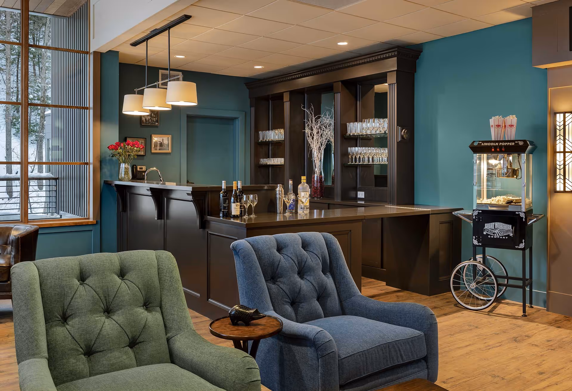 A cozy lounge area with two upholstered armchairs, one green and one blue, facing a dark wood bar counter with bottles and glasses. Behind the bar is a mirrored shelving unit with glassware. To the right, there is a vintage-style popcorn machine on a stand. Large windows on the left show a snowy outdoor scene.