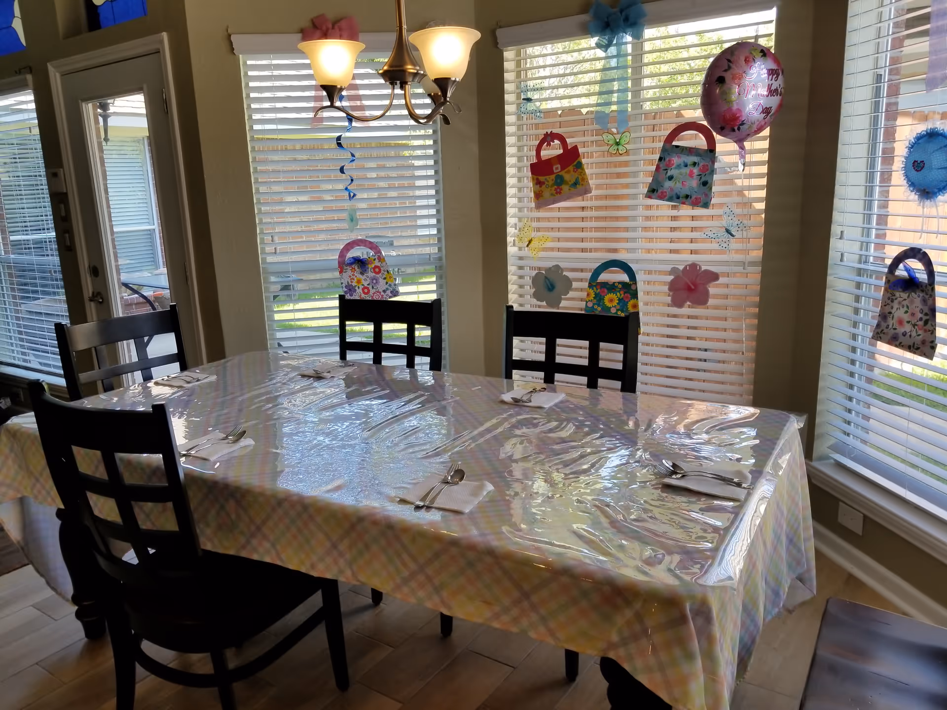Dining room with a rectangular table covered with a pastel plaid tablecloth and a clear plastic cover. The table is set with napkins and silverware for six people. Four black wooden chairs surround the table. Large windows with white blinds let in natural light, and colorful paper decorations including handbags, flowers, butterflies, and a 'Happy Mother's Day' balloon are attached to the windows. A chandelier with three lights hangs above the table.