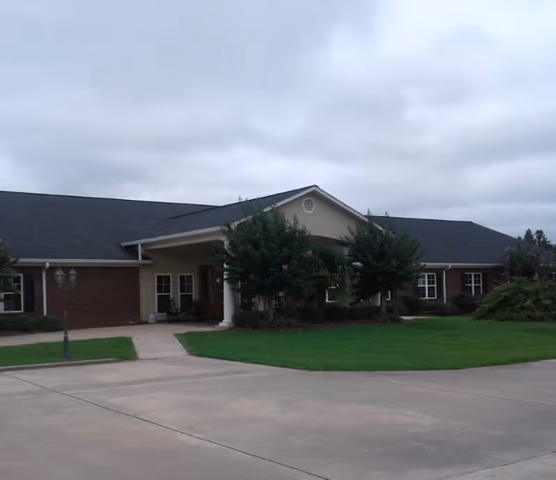 Exterior view of a single-story senior living facility building with a covered entrance supported by white columns, surrounded by green lawn and small trees under a cloudy sky.