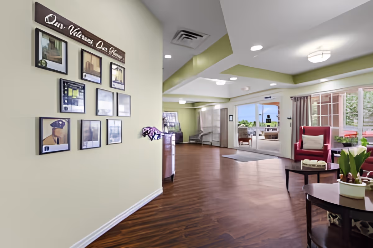 Interior view of a senior living facility lounge area with wooden flooring, light green walls, framed pictures on the wall, red armchairs with cushions, a coffee table with a plant, and large windows and glass doors leading to an outdoor patio area.