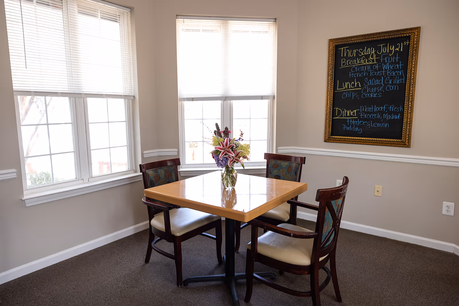 A small dining area with a square wooden table and four chairs with patterned upholstery. A vase with a colorful flower arrangement is placed in the center of the table. Two large windows with white blinds allow natural light into the room. On the wall to the right, there is a framed chalkboard displaying the menu for Thursday, July 21st, listing breakfast, lunch, and dinner options.
