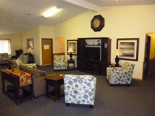 A common area in a senior living facility featuring several upholstered armchairs with floral patterns, a sofa, wooden side tables, a large dark wood entertainment center with a television, two table lamps, framed artwork on the walls, and a clock above the entertainment center. The room has carpeted flooring and neutral-colored walls.