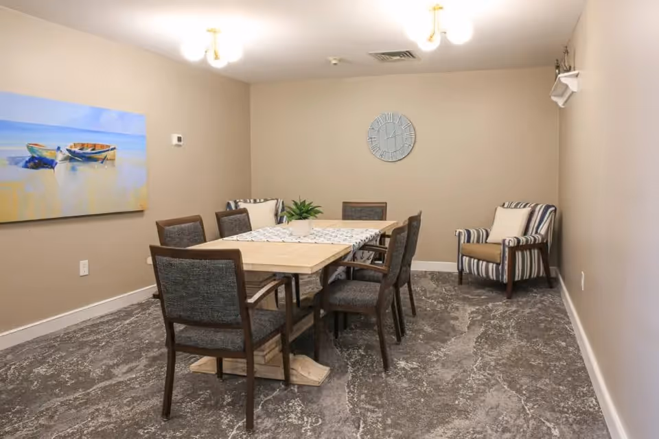 A dining room with a rectangular wooden table surrounded by six chairs with dark upholstery. There is a small plant centerpiece on the table and a decorative runner. The room has beige walls, a gray patterned carpet, a wall clock, a painting of two boats on the left wall, and a striped armchair with a cushion in the corner.