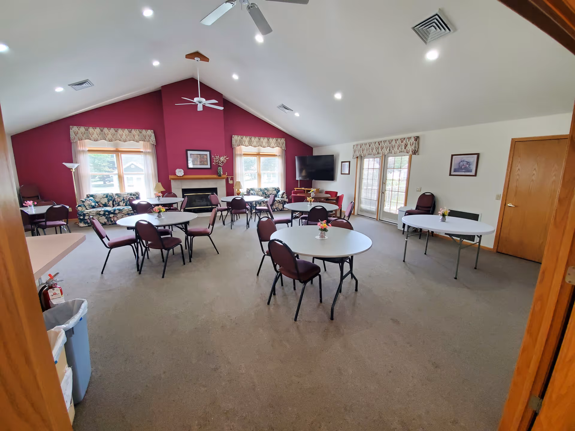 A spacious common area with several round tables and chairs arranged throughout the room. The room features a high vaulted ceiling with ceiling fans and recessed lighting. There are two large windows with floral valances on a deep red accent wall, a fireplace beneath a framed picture, and two floral-patterned sofas. A large flat-screen TV is mounted on the right wall near glass double doors. The room has beige carpeting and light-colored walls with framed artwork.