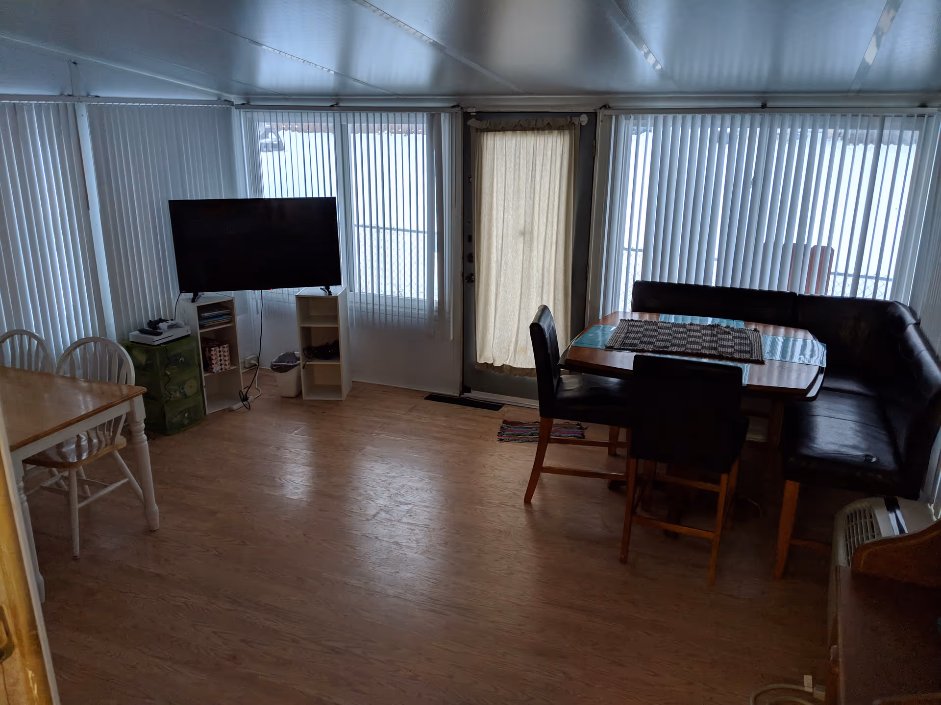 Interior room with wooden flooring featuring a dining table with black chairs and a corner bench, a flat-screen TV mounted on a small white shelf unit, a wooden table with white chairs, and large windows with vertical blinds letting in natural light.