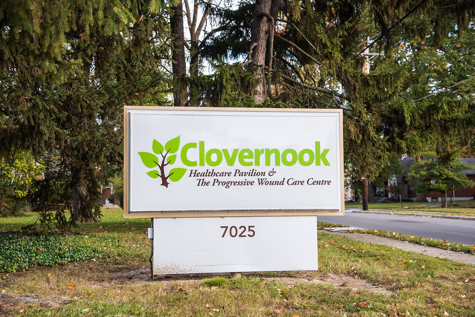Outdoor sign for Clovernook Healthcare Pavilion & The Progressive Wound Care Centre with the address number 7025, surrounded by trees and grass near a street.