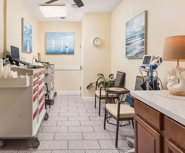 Narrow interior medical/treatment room with supply carts on wheels, chairs, medical equipment, wall art and a clock on pale yellow walls.