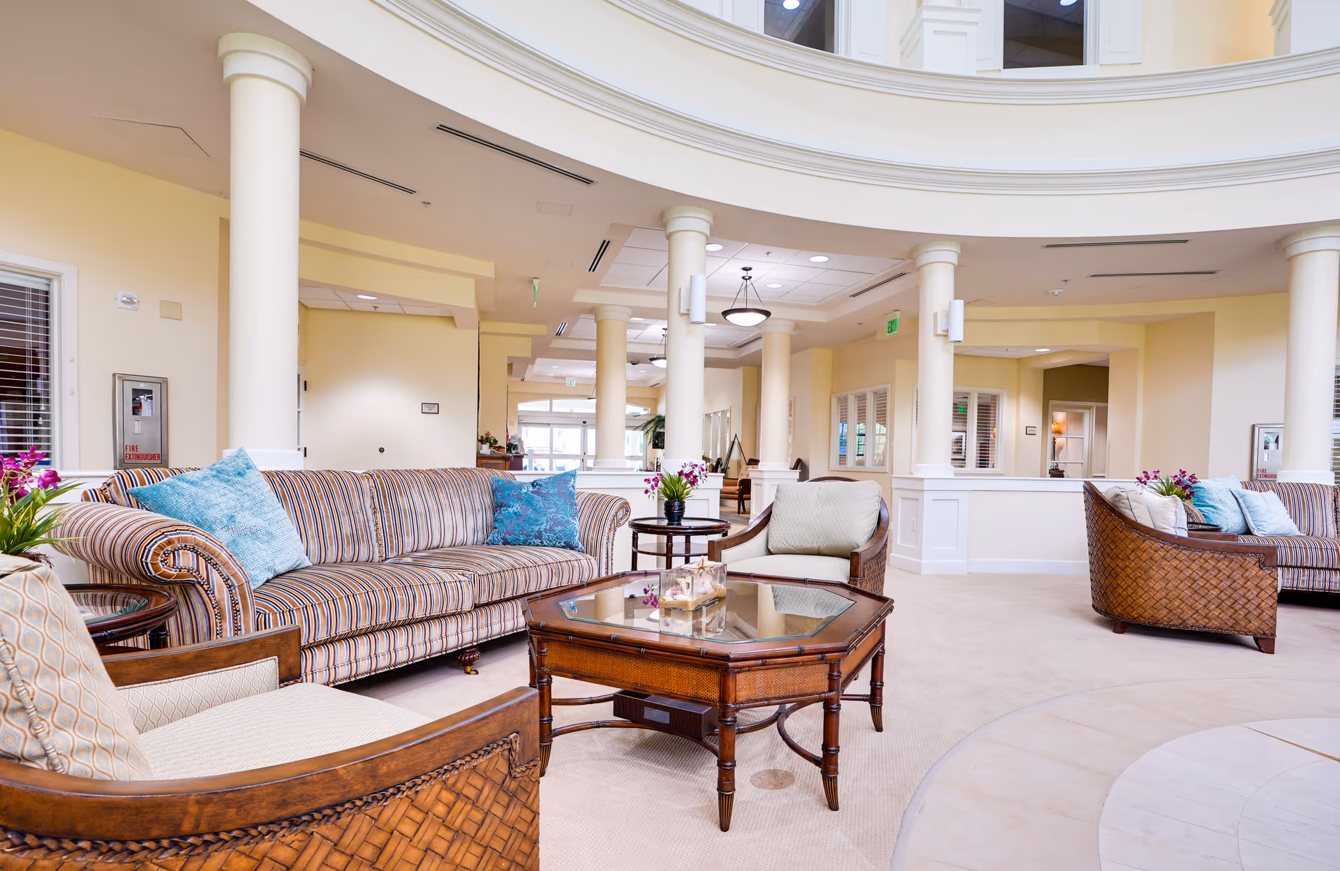 A spacious and well-lit living room area in a senior living facility featuring striped and wicker sofas with blue and beige cushions, a wooden coffee table with glass top, beige carpet, cream-colored walls, white columns, and ceiling lights.