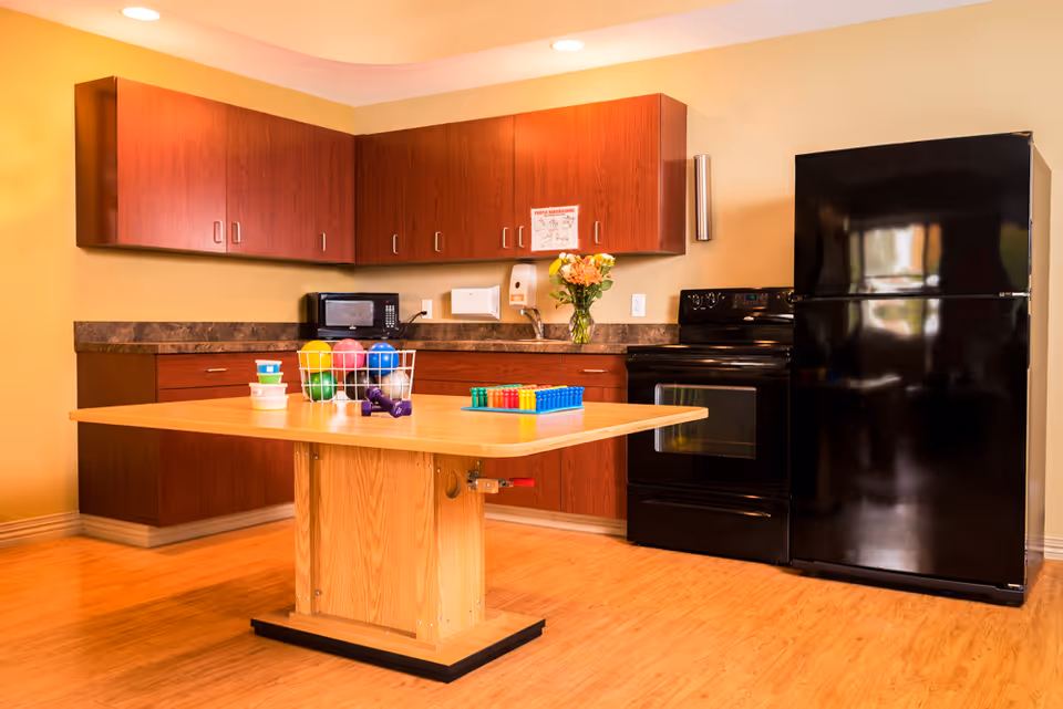 A kitchen area with wooden cabinets, a black refrigerator, a black stove, a microwave, and a wooden table in the center. The table has colorful exercise balls, small dumbbells, and other small items on it. The floor is wooden, and there is a vase with flowers on the countertop.