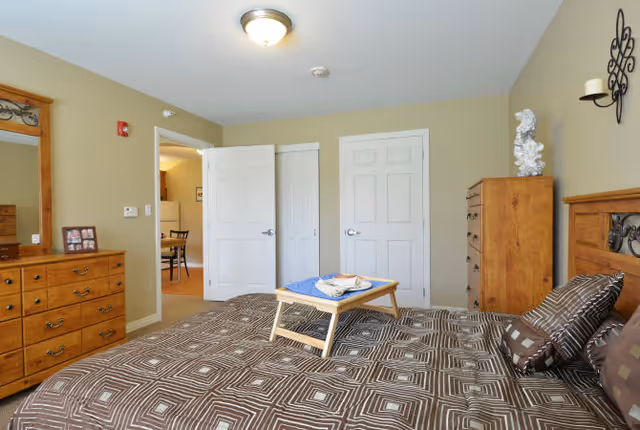 A bedroom with a large bed covered in a brown geometric patterned bedspread and matching pillows. A wooden tray with a blue cloth and some items is placed on the bed. The room has beige walls, a wooden dresser with a mirror, a tall wooden chest of drawers, and two white doors. A small wall-mounted candle holder and decorative piece are visible on the wall above the chest of drawers. The doorway leads to another room with a dining table and chairs.