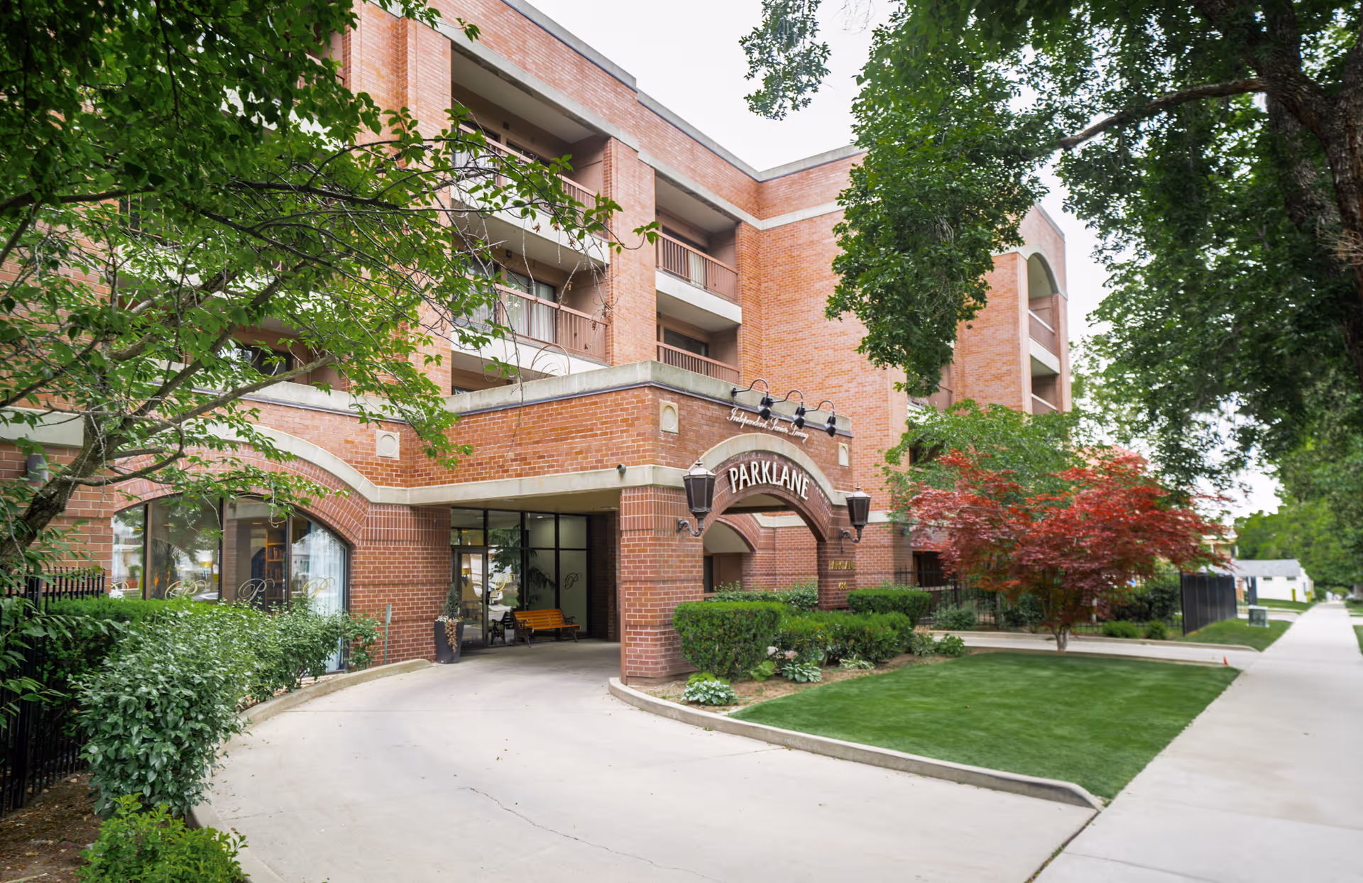 Exterior view of Park Lane Senior Living facility showing a brick building with multiple balconies, an arched entrance with the name 'Park Lane' displayed, surrounded by green trees, bushes, and a well-maintained lawn.