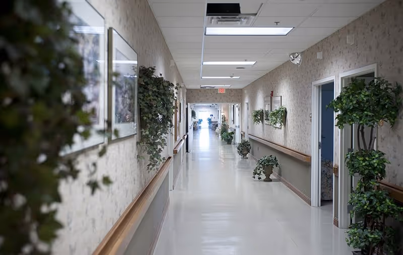 A long, well-lit hallway in a senior living facility with beige walls decorated with framed pictures and green plants. The floor is light-colored and shiny, with handrails on both sides. Several doorways lead to rooms along the corridor, and there is an exit sign visible at the far end.