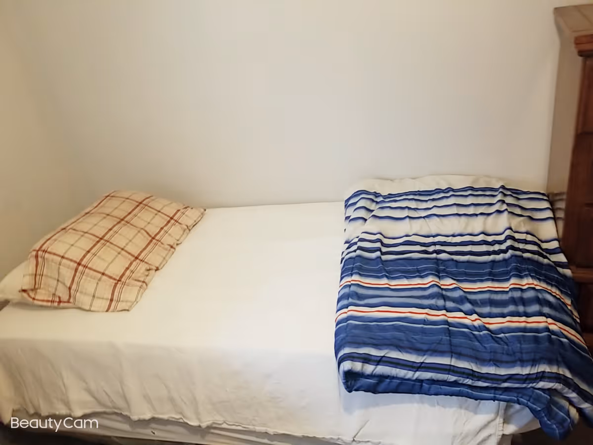 A simple bedroom with a single bed against a plain wall. The bed has a white sheet, a plaid pillow, and a blue striped blanket partially covering it. There is a wooden piece of furniture visible on the right side.