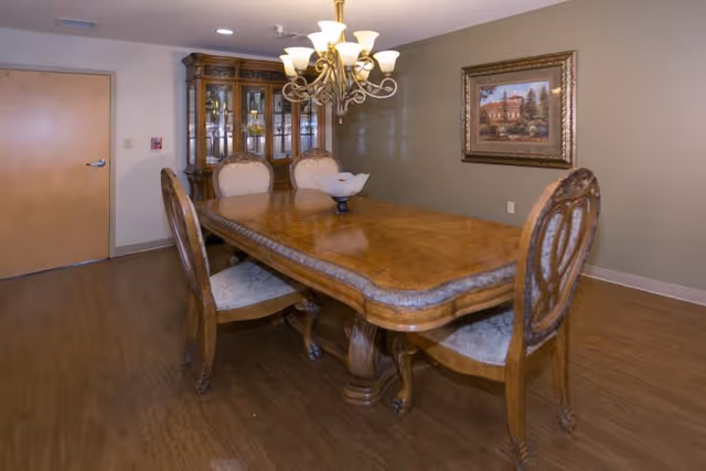 A dining room with a large wooden dining table and six upholstered chairs. A chandelier with multiple lights hangs above the table. There is a wooden china cabinet with glass doors in the corner and a framed painting on the wall. The floor is wood and there is a closed wooden door on the left side.