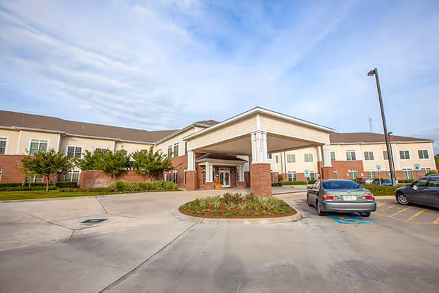 Front exterior view of a two-story senior living facility with a covered entrance, brick and beige siding, landscaped greenery, and a parking area with cars.