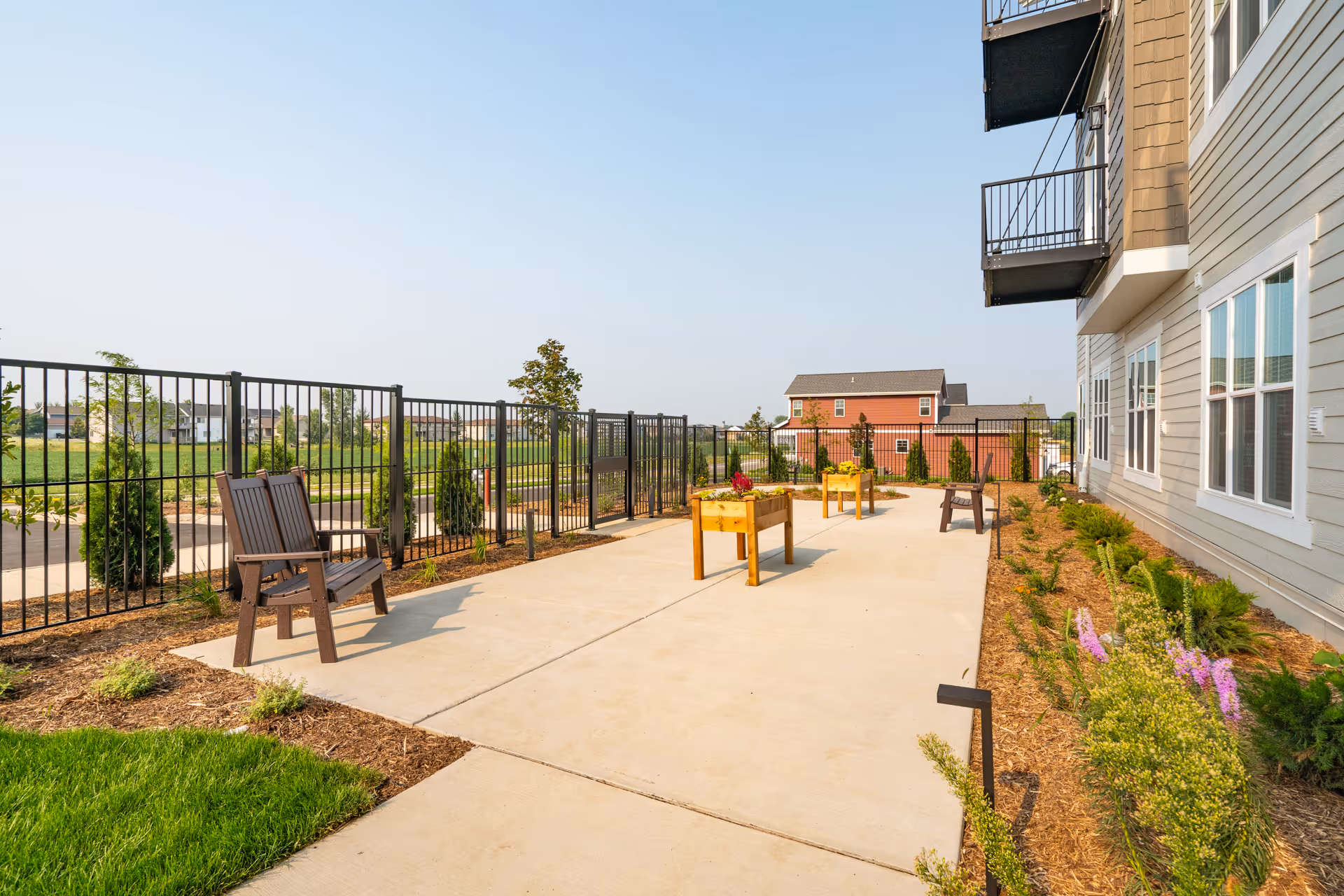 Outdoor patio area at Talamore Senior Living Sun Prairie with wooden chairs, raised garden beds, and a fenced perimeter. The patio is adjacent to a building with multiple windows and balconies, surrounded by landscaped greenery and plants under a clear sky.