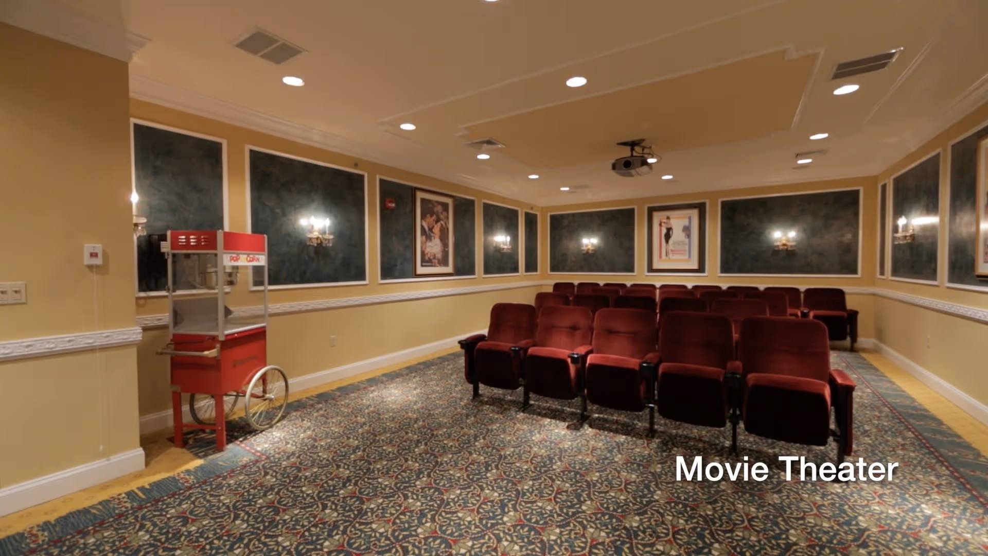 Interior of a small movie theater room with rows of red cushioned seats, a popcorn machine on the left side, decorative wall sconces, framed movie posters on the walls, and a patterned carpeted floor.