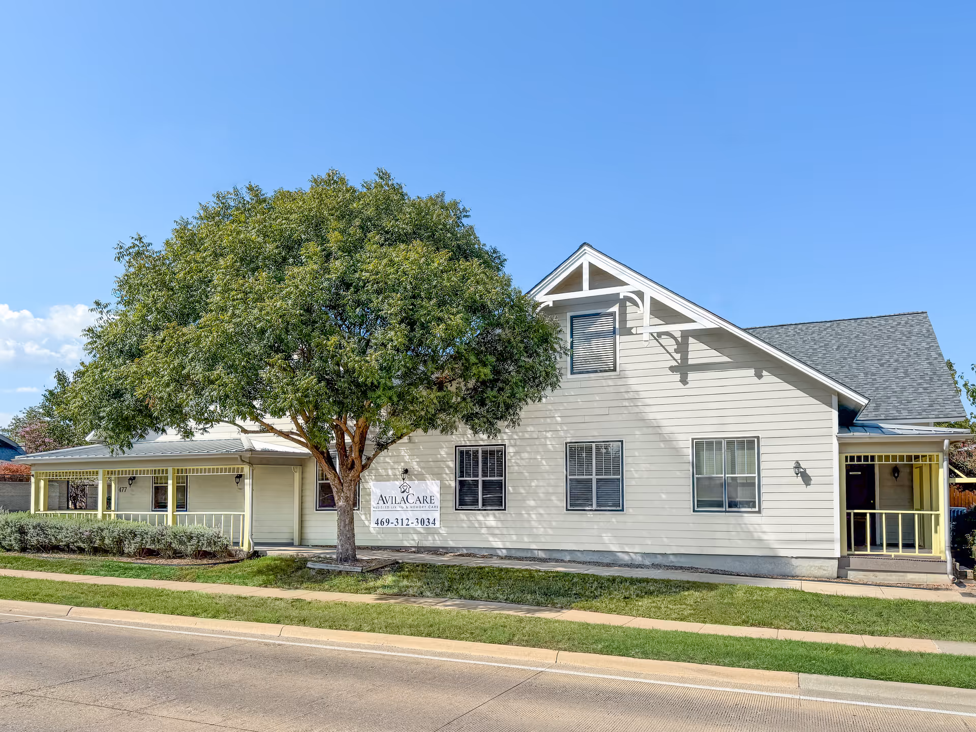 Exterior view of a single-story light-colored building with a large tree in front and a sign that reads 'AvilaCare Assisted Living & Memory Care 469-312-3034'. The building has multiple windows and a covered porch area. The sky is clear and blue.